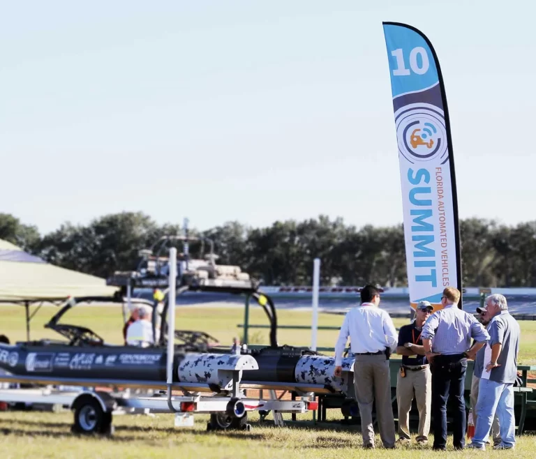 A group of people gathered outside at a demonstration area during the Florida Automated Vehicles conference, marked by a large 12' branded banner.